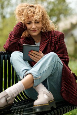Rahat bir kadın, ayakları üzerinde bir bankta oturuyor ve e-kitap okuyor. Onun saçı kırmızı ve kıvırcık. Kırmızı ekose ceket, kot pantolon ve ayakkabılar. Eski hobilerin tadını çıkarıyorum..