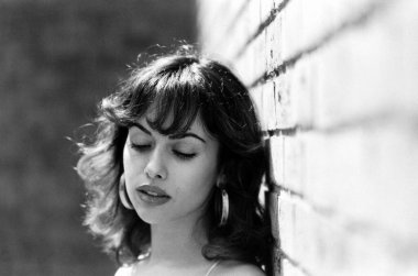 Tuğla duvara yaslanan genç model kadının tek renkli yakın portresi. Güneşli bir gün ve ışık arkadan geliyor. Kocaman küpeler takıyor. Film analog kamerasıyla yapılmış tahıllı resim.