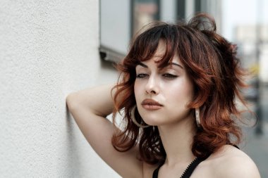 Beyaz bir duvara dayanmış genç bir kadının nostaljik portresi. Saçları kırmızı ve sağ dirseği duvarda. Başka tarafa bakıyor..