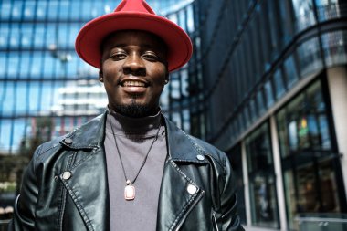 Kırmızı şapkalı gülümseyen adam dışarıdaki bir iş ortamında kameraya bakıyor. Arkasında modern cam bir bina var. Deri ceket giyiyor..