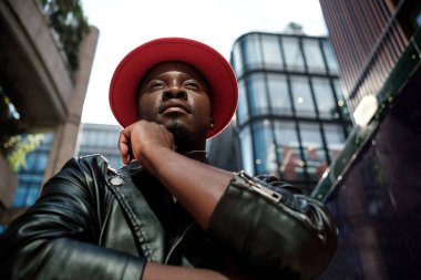 Bir adam arkasında yüksek binalar olan ciddi bir ifadeyle poz veriyor. Cam binalarla dolu bir iş alanında. Kırmızı şapka ve siyah deri ceket giyiyor..