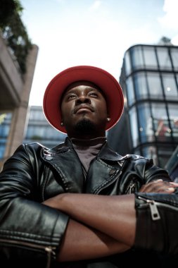Modern binaların önünde kollarını kavuşturarak poz veren gururlu erkek model. O, kırmızı bir şapka ve siyah deri ceket giyiyor. Kendine güvenini ve kişiliğini gösteriyor..