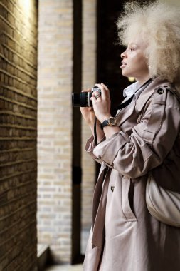 Albino kadın elinde analog bir kamera tutuyor ve başka tarafa bakıyor. O bir trençkot giyiyor ve saçları sarı ve kıvırcık. Dijital çağda geleneksel sanatın tadını çıkarmak.