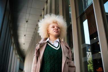 Albino model kadın, modern, geniş bir koridorda poz veriyor. Açık havada bir sürü sütun var. Altında bir trençkot ve yeşil örgü bir yelek var. Saçları kıvırcık ve sarı..