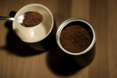 Metal kaşık, boş bir fincanın üstündeki hazır kahve tanelerini kaldırıyor. Tahta bir masada, sıcak suyla karıştırılmaya hazır bir kahve kutusunun yanında.