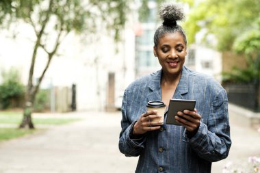 Parktaki olgun bir kadın e-kitap kullanıyor ve elinde tek kullanımlık bir fincan kahve tutuyor. Gülümsüyor ve okumaktan zevk alıyor..