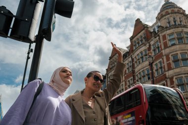Caddede taksi çağıran iki bayan arkadaş. Arkasında klasik bir Londra kırmızı otobüsü var. Yoğun şehir sokakları konseptinde işe gidip gelmek. Çok kültürlü arkadaşlık kavramı.