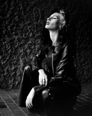 Rahatlamış genç bir kadın dışarıda poz veriyor. O, gözleri kapalı kaba duvarın önünde. O, siyah deri kıyafetler giyiyor. Tek renkli analog resim.