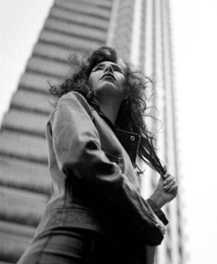 Gökdelenin önündeki genç bayana düşük bir bakış açısı. O başka yere bakıyor ve siyah bir deri ceket giyiyor. Alternatif görünüş. Monokrom analog film görüntüsü.