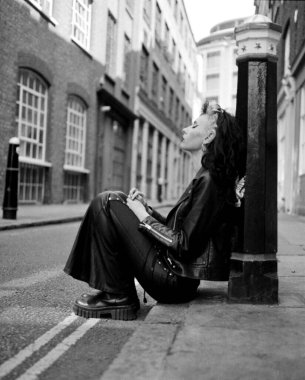 Alternatif olarak genç bir kadın yerde dinleniyor ve caddede bir kordona yaslanıyor. O, siyah bir deri elbise giyiyor. Tek renkli analog resim.
