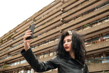 Dişi rock yıldızı eski binanın önünde selfie çekiyor. Siyah deri bir ceket giyiyor ve saçı dağınık. Teknoloji ve iletişim konsepti.
