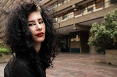 Geniş açılı objektifle yapılmış alternatif genç bir kadının vesikalığı. Arkasında bulanık bir bina var. Saçları dağınık ve dağınık. Makyajı çok ağır..