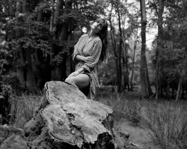 Uzun saçlı, desenli bir elbise giyen, ormanda büyük bir ağaç kütüğünde oturan, doğanın sükunetini kucaklayan genç bir kadının tek renkli orta formatlı fotoğrafı.
