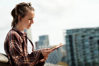 Şık saç kesimi ve piercingleri olan genç bir kadın bir kitap okumaya dalmış, çağdaş şehir mimarisinin arka planına karşı bir bankta oturuyor.