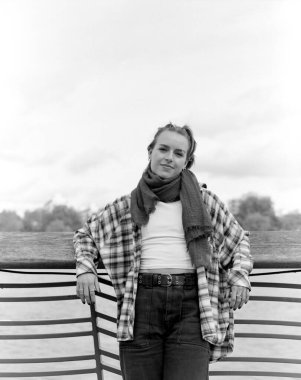 Renkli tek renkli orta sınıf bir film portresi. Atkısı ve kareli gömleği olan, parmaklıklara yaslanmış, bulutlu bir gökyüzü ve bulanık bir arkaplan manzarasının keyfini çıkaran zarif bir genç kadın.