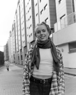 Ekose ve eşarp takmış gülümseyen genç bir kadının arka planda binaları olan bir şehir sokağında dikilirken tek renkli orta formatlı film portresi.