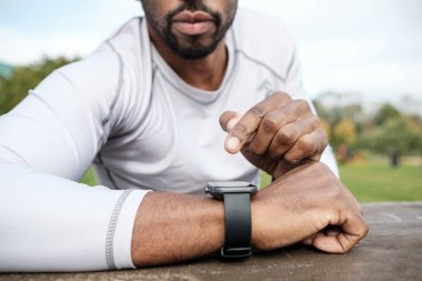 Uzun kollu beyaz gömlek giyen sporcu, parkta bir eğitim molası sırasında akıllı saatinin verilerini kontrol ediyor ve performansını izlemek için teknolojiyi kullanıyor.
