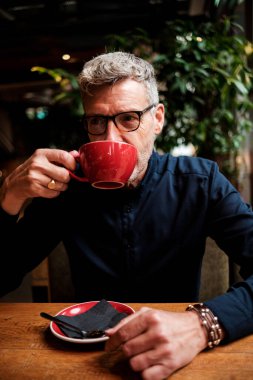 Gri saçlı iş adamı, gözlük ve mücevher takıyor modaya uygun bir kafede kahve içiyor, ahşap bir masada otururken kırmızı bardaktan bir yudum alıyor.
