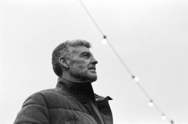 Kışlık ceket giymiş, arka planda bulanık sicim ışıklarıyla yukarı bakan olgun, gri saçlı bir adamın tek renkli analog fotoğrafı.