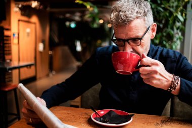 Olgun iş adamı kırmızı bir fincan kahveden içiyor ve kafede gazete okuyor, iş günü boyunca sakin bir anın tadını çıkarıyor.