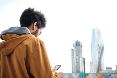 Afro saçlı, kahverengi kapüşonlu ceket giyen, arka planda modern gökdelenlerle dolu bir şehir ortamında akıllı telefon kullanan genç adam, şehir yaşamında teknoloji ve iletişimi somutlaştırıyor.