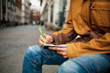 Titiz bir şekilde not defterine yeşil kalemle not alan, bulanık bir şehir ortamında dışarıda oturan, bir haber ya da makale için ayrıntıları yakalayan bir gazetecinin yakın çekimi.