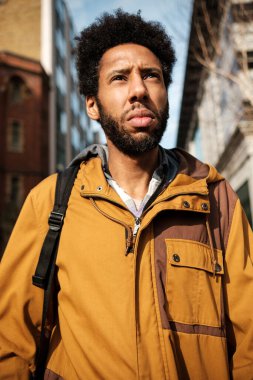 Sıradan bir ceket giyen ve sırt çantası taşıyan genç bir adamın portresi, parlak, güneşli bir gökyüzünün altında bir şehir caddesinde dikilirken dikkatlice yukarı bakıyor.