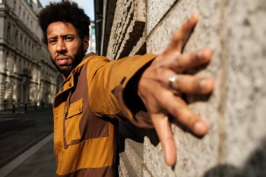 Şık kahverengi ceket giyen genç, modaya uygun bir adam, canlı bir şehir caddesinde duvara yaslanırken elini kameraya doğru uzatarak güven içinde poz veriyor.