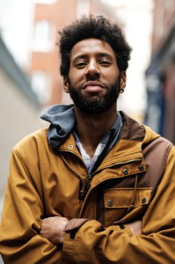 Afro saç stili ve çapraz kollu Afro Amerikalı genç adam şehir ortamında kendine güvenen bir poz veriyor, şık bir mermer ceket giyiyor bulanık şehir arka planına karşı