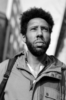 Siyah beyaz, analog bir film. Afro saçlı, günlük giysiler giymiş ve sırt çantasıyla yukarı bakan genç bir adam. Düşünce ya da arzu hissi veriyor.