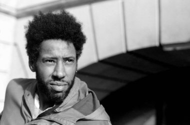 Düşünceli bir ifadeye ve afro saç stiline sahip genç bir adam, kentin arka planına karşı poz veriyor, tek renkli, klasik 35 mm 'lik bir filmi anımsatıyor.
