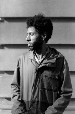 Grainy monokrom analog vinog fotografta kapüşonlu ceket giyen, duvarın yanında duran, 35 mm 'lik bir filme çekilen Afro saçlı bir genç resmediliyor.