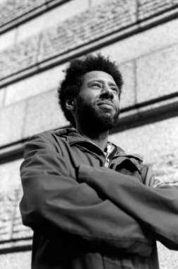 Kolları katlanmış, taş bir duvara bakan, 35 mm 'lik bir kamerayla yakalanmış, Afro-Amerikan bir adamın tek renkli ve klasik bir fotoğrafı.