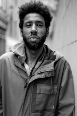 Kapüşonlu ceket giyen ve şehir duvarına yaslanmış, klasik bir film estetiğine bürünmüş, şık bir genç adamın siyah beyaz portresi.