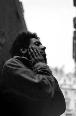 Tahıllı tek renkli analog fotoğraf dalgın, kıvırcık saçlı, iki eliyle yüzünü tutan bir adam görüntüsü bulanık şehir binalarının arka planında kaybolmuş gibi görünüyor.