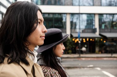 Genç, şık bir çift şehir caddesinde duruyor, uzaktaki bir şeye bakıyor, modern binalar ve arka planda bulanık bir restoran var.