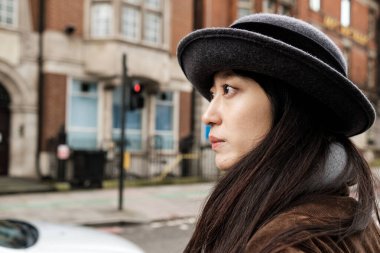 Şapkalı ve kahverengi kadife ceketli genç Asyalı kadın Londra 'nın bir caddesinde yürüyor, ciddi bir ifadeyle sağına bakıyor.