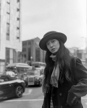 Kasvetli siyah beyaz bir fotoğraf şapkalı ve atkılı şık bir kadını çekiyor, şehir ortamında bulanık arabalarla dikiliyor, zamansız bir klasik atmosferi çağrıştırıyor.