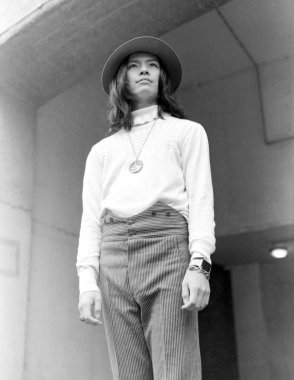 1970 'lerin modasında genç bir adamı gösteren taneli tek renkli bir fotoğraf. Şapkası, kolyesi olan bir süveteri ve beton bir duvara dayanmış yüksek bel çizgili pantolonu var.