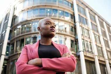 Kollarını kavuşturmuş bir iş kadını modern bir ofis binasının önünde duruyor ve iş dünyasına güven ve başarı aktarıyor.