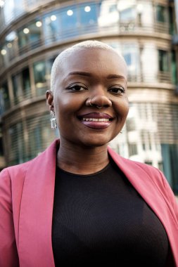 Modern bir ofis binasının önünde gülümseyen, iş dünyasında başarılı bir kadın liderliği temsil eden kısa boyalı saçları ve burun piercingli kendine güvenen bir iş kadını.