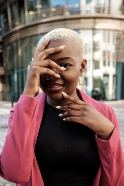 Kısa sarı saçlı şık bir iş kadını, pembe bir ceket giyiyor, yüzünün bir kısmını eliyle kapatıyor, modern bir ofis binasının yanında poz veriyor, özgüven ve profesyonellik saçıyor.