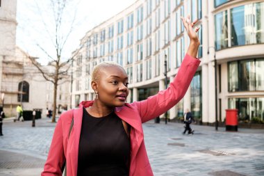 İngiltere, Londra 'da taksi çağıran pembe ceketli genç bir iş kadını modern ofis binalarının yanındaki kaldırımda yürürken...