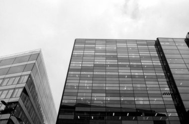 Modern cam binalar bulutlu bir gökyüzüne karşı dimdik duruyorlar, klasik, kumlu tek renkli film estetiği ile çekiliyor, kentsel manzaraya zamansız bir kalite katıyor.