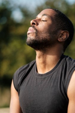 Genç erkek sporcu derin bir nefes alıp temiz havanın tadını çıkarıyor ve huzurlu bir park ortamında zihinsel ve fiziksel refahı teşvik ediyor.