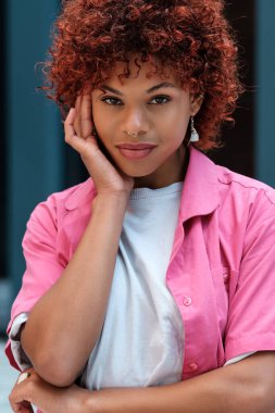 Kıvırcık saçlı, burnu halkalı, kendine güveni tam genç bir kadın. Düşünceli bir poz veriyor. Pembe gömleği ve tarzı var.