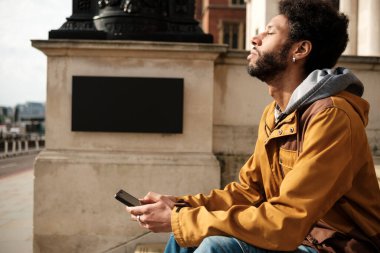 Genç siyahi adam, gözleri kapalı meditasyon yapıyor ve elinde bir akıllı telefon tutuyor. Londra 'daki Trafalgar Meydanı' nın önünde oturuyor. Bir anlık farkındalığın tadını çıkarıyor.