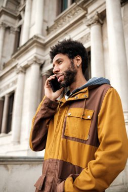 Afro saçlı genç bir adam telefon görüşmesi yapıyor. Sütunları olan tarihi bir binanın önünde duruyor. Günlük kahverengi ve sarı bir ceket giyiyor.