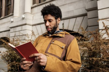 Bir üniversite binasının önünde duran genç üniversite öğrencisi, bir kitaba dalmış, yaklaşan sınavlara hazırlanırken bilgiyi özümsüyor ve akademik atmosferin tadını çıkarıyor.