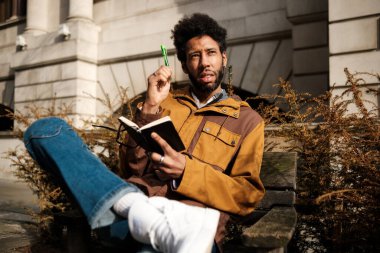 Genç yazar, elinde bir defter ve kalemle binanın dışındaki bankta oturuyor. Bir sonraki eseri için ilham toplarken düşünceli görünüyor.